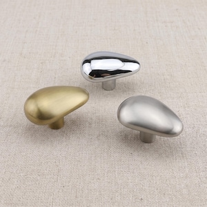 May include: Three cabinet knobs with an organic, pebble-like shape. One knob has a polished chrome finish, one has a brushed gold finish, and one has a brushed silver finish. Each knob features a smooth, rounded top and a central mounting post.