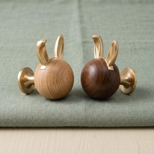 May include: Two spherical wall hooks featuring rabbit ears made of brushed gold-tone metal. One hook has a light-toned natural wood sphere, while the other has a dark-toned wood sphere. Each hook includes a circular gold-tone mounting base.