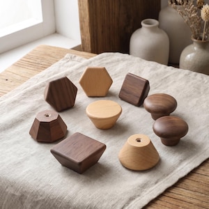 Puede incluir: Una colección de nueve pomos de madera para armarios en diversas formas y tonos de madera. El conjunto incluye diseños hexagonales, cuadrados, redondos y cónicos en acabados de haya clara y nogal oscuro, cada uno con un orificio central de metal.
