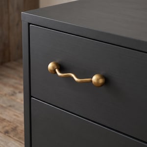 Puede incluir: Una cómoda de madera en color negro mate con un cajón horizontal. El cajón cuenta con un distintivo tirador de metal en tono latón que presenta una forma ondulada y sinuosa, terminando en dos extremos esféricos y lisos.