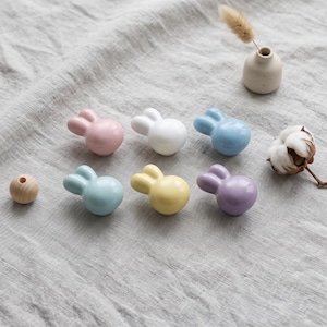 May include: A set of six ceramic cabinet knobs shaped like rabbit heads in pastel pink, white, light blue, teal, yellow, and lavender, accompanied by a single small wooden bead, a small ceramic vase, and a dried cotton flower stem.