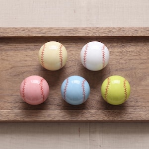 May include: Five spherical knobs shaped like baseballs, each featuring red stitching details. The set includes one cream, one white, one light pink, one light blue, and one lime green knob, all arranged in a cluster on a flat wooden surface.