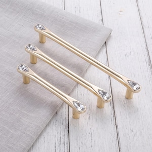 May include: Three gold-toned metal cabinet pulls of varying lengths, each featuring a smooth, cylindrical bar design. Every pull is accented with a clear, faceted, teardrop-shaped crystal inlay at both ends, secured by two small cylindrical posts.