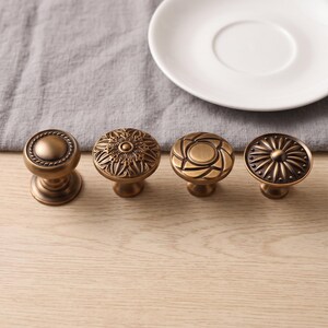 May include: Four round cabinet knobs with an antique brass finish. The first knob has a smooth center with a rope-textured border. The second features an intricate floral pattern. The third has a geometric swirl design. The fourth has a sunburst pattern.