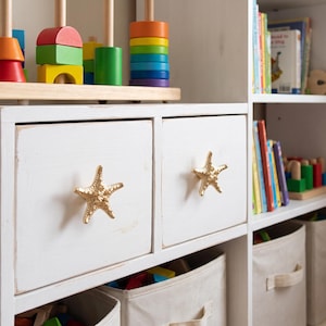 May include: A white wooden storage unit featuring two square drawers with gold-colored starfish-shaped knobs. The unit includes open shelving filled with colorful wooden stacking toys and various books, alongside beige fabric storage bins below.