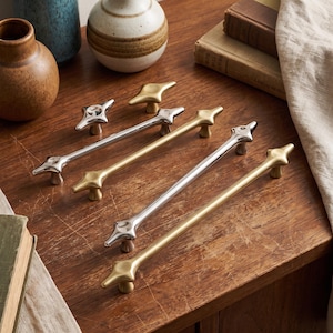 May include: A collection of four elongated metal cabinet pulls arranged diagonally. Two pulls feature a polished silver finish, while the other two have a matte gold finish. Each pull has a central bar with star-shaped ends.
