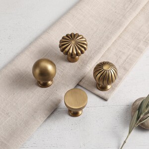 May include: Four brass-toned cabinet knobs with a brushed metallic finish. One knob is a smooth sphere, one is a flat-topped cylinder, and two feature a radial ribbed pattern with a flower-like design. Each knob has a circular base.