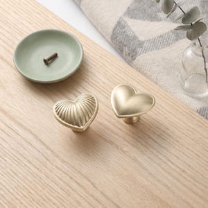 May include: Two heart-shaped cabinet knobs with a matte gold finish. One knob features a ribbed, textured surface, while the other has a smooth, plain surface. Both knobs are designed for furniture attachment and have a rounded, dimensional shape.