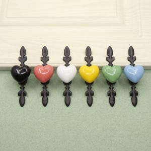 Könnte beinhalten: Ein Set aus sechs Schubladengriffen, jeweils mit einem herzförmigen Knopf in verschiedenen Farben: Schwarz, Rosa, Weiß, Gelb, Grün und Blau. Jedes Herz ist auf einer vertikalen schwarzen Metallplatte mit blattartigem Design montiert.