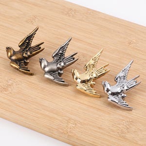 Puede incluir: Cuatro pomos de cajón o ganchos de pared de metal con forma de pájaro dispuestos en fila. El conjunto incluye un acabado en latón antiguo, gris oscuro mate, oro pulido y plata pulida, cada uno representando un pájaro en pleno vuelo.