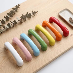 May include: A set of six arched cabinet handles in a glossy finish, arranged in a diagonal row. The colors include white, light pink, light blue, green, yellow, orange, and red. Each handle features a smooth, rounded, U-shaped design.