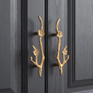May include: Two gold-colored metal cabinet pulls shaped like tree branches with textured bark and small floral accents. Each handle features two distinct flower blossoms attached to the branch structure, mounted vertically on dark gray cabinet doors.