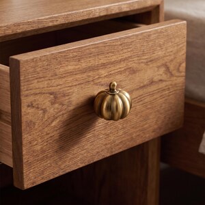 May include: A brass-colored metal drawer knob shaped like a small pumpkin with vertical ridges, attached to the front of a light brown wooden drawer that is partially pulled open, revealing the natural wood grain texture of the furniture.