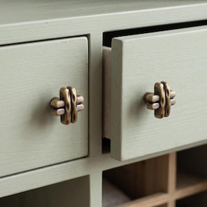 May include: A sage green wooden cabinet featuring two drawers with antique brass-toned metal knobs. The knobs have a textured, ribbed design with a central vertical bar. One drawer is partially open, revealing a light-colored interior surface.