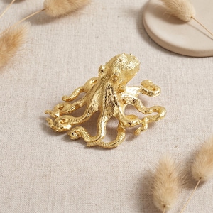 May include: A gold-toned octopus brooch featuring a textured, metallic surface with intricate detailing on the head and multiple curved tentacles extending outward, creating a realistic representation of the marine creature in a polished finish.