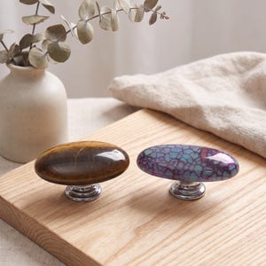 May include: Two oval-shaped cabinet knobs with polished silver-tone metal bases. One knob features a smooth, golden-brown tiger's eye stone pattern, while the other displays a vibrant purple and teal crackled stone texture with a glossy finish.