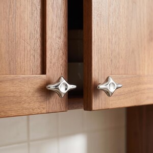 Puede incluir: Dos pomos de armario de cromo pulido con forma de estrella de cuatro puntas fijados a puertas de armario de madera de color marrón claro. Los pomos presentan un acabado metálico liso y reflectante con una hendidura circular central.