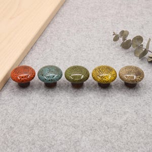 May include: Five round ceramic cabinet knobs arranged in a row, each featuring a unique speckled glaze in orange, teal, olive green, yellow, and beige. These mushroom-shaped drawer pulls have a glossy finish and a dark brown base.