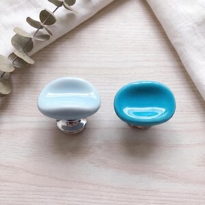 May include: Two rounded cabinet knobs with a smooth, glossy finish. One knob is a pale, soft blue, while the other is a vibrant, saturated turquoise. Both feature a polished silver-toned metal base that connects to the rounded top surface.