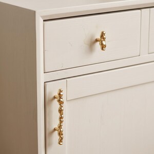 May include: A light-colored wooden cabinet featuring a single drawer and a door. The drawer is fitted with a small, irregular, gold-toned knob, while the door includes a long, vertical, gold-toned handle with a textured, organic, molten-like shape.