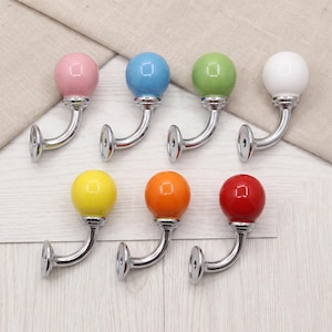 May include: A set of seven wall hooks, each featuring a smooth, spherical knob in a different solid colour: pink, blue, green, white, yellow, orange, and red. Each hook has a polished silver-tone metal arm and a circular base with a screw hole.