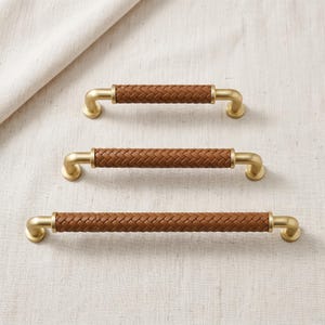 May include: Three cabinet pulls arranged vertically, each featuring a central brown leather-wrapped handle with a woven texture and brushed gold metal ends. The pulls vary in length, with the shortest at the top and the longest at the bottom.