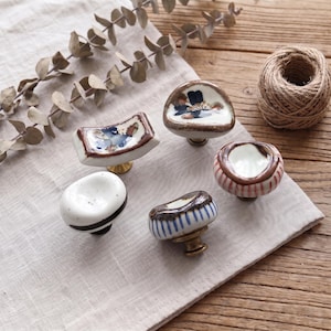May include: A collection of five ceramic cabinet knobs with varied shapes and hand-painted designs. The set includes pieces with blue floral patterns, white surfaces with dark speckles, and vertical stripes in blue or red, all featuring metallic bases.