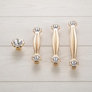 May include: A collection of gold-toned cabinet hardware including one round knob and three varying lengths of drawer pulls. Each piece features a polished metallic finish and is accented with clear, faceted circular crystals at the ends.