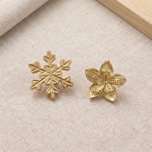 Könnte beinhalten: Zwei goldfarbene Metallanhänger, einer in Form einer sechsstrahligen Schneeflocke mit symmetrischen geometrischen Zweigen und der andere in Form einer fünfblättrigen Blume mit strukturierten, geäderten Blütenblättern, die von einem Mittelpunkt ausgehen.