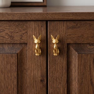 May include: Two gold-colored metal cabinet knobs shaped like sitting foxes with pointed ears and detailed facial features. These metallic animal-shaped pulls are mounted onto the vertical wooden panels of a dark brown cabinet door set.
