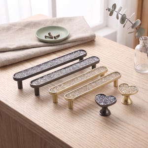 May include: A collection of cabinet hardware including two long dark gray rectangular pulls, two long gold rectangular pulls, one dark gray heart-shaped knob, and one gold heart-shaped knob, all featuring a textured, clear crystal-like inlay.
