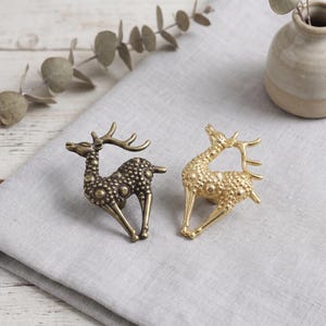 Puede incluir: Dos pomos de metal para muebles con forma de ciervo, uno con acabado en bronce oscuro y otro en dorado brillante. Cada figura de ciervo presenta un cuerpo texturizado con detalles circulares en relieve, patas delgadas y astas curvadas.