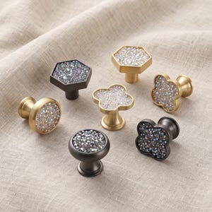Puede incluir: Una colección de siete pomos para armarios en varias formas, incluyendo diseños redondos, hexagonales y de trébol. Cada pomo presenta una base metálica en tonos dorados o gris oscuro, con una incrustación central de pequeños cristales brillantes.