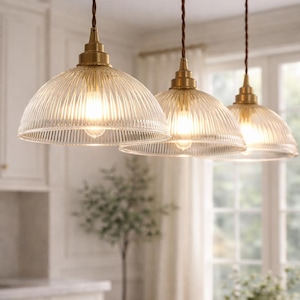 May include: Three pendant lights with clear, ribbed glass shades and brass-colored hardware. The lights hang from brown twisted cords. The kitchen setting has a neutral color palette with a marble countertop and a window in the background.