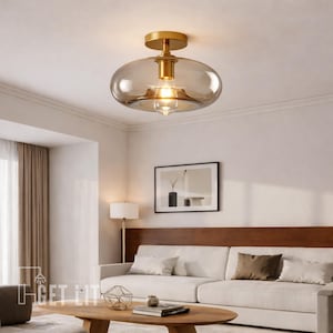 Könnte beinhalten: Eine moderne Deckenleuchte mit goldfarbenem Sockel und einem transparenten, oval geformten Glasschirm. Das Licht ist eingeschaltet und beleuchtet den Raum. Die Leuchte ist an einer weißen Decke montiert. Der Raum hat ein Sofa, einen Couchtisch und Kunstwerke.