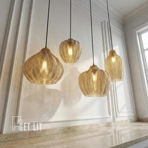 May include: Four pendant lights with textured, amber-colored glass shades hang from black cords. The shades have a woven pattern and are illuminated from within. The lights are suspended against a white wall with decorative molding, above a light-colored countertop.