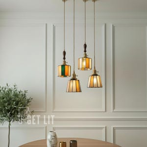 Könnte beinhalten: Vier Pendelleuchten mit Buntglasschirmen in verschiedenen Farben, darunter Grün, Orange und Creme. Jede Leuchte hat eine Messingarmatur, einen Holzgriff und ein langes Kabel. Die Leuchten hängen von der Decke.