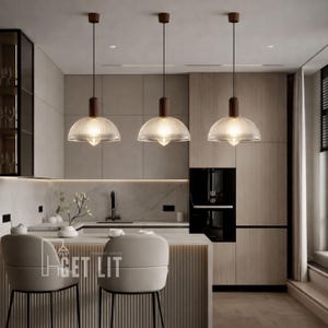 May include: Three pendant lights with clear, ribbed glass shades and dark brown wooden accents hang above a kitchen island. The kitchen features light wood cabinetry, a marble countertop, and two bar stools.
