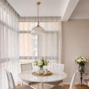 May include: A dining room with a white round table, four white chairs, and sheer curtains. A glass pendant light hangs above the table. A vase of white flowers sits on the table, and another vase of white flowers is on a decorative stand.