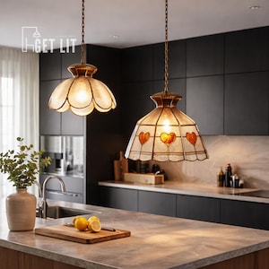 May include: Two pendant lights with stained glass shades hang above a kitchen countertop. One shade has a scalloped edge, the other has heart-shaped designs. The lights are suspended by gold-coloured chains. The kitchen has dark cabinets and a marble countertop.