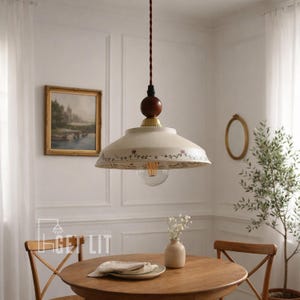 Puede incluir: Lámpara colgante de color crema con un diseño floral alrededor del borde, colgando sobre una mesa de madera. La lámpara tiene una bombilla transparente y un cable marrón oscuro. La habitación cuenta con una pintura, un espejo y un pequeño jarrón con flores.