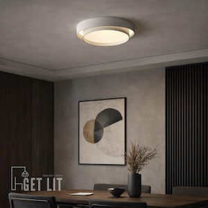 Op de afbeelding: Een moderne, ronde plafondlamp met een witte buitenring en een warm, gloeiend binnenlicht. De armatuur is gemonteerd op een neutraal gekleurd plafond. De afbeelding toont ook een eetkamer met een tafel, stoelen en kunstwerk.