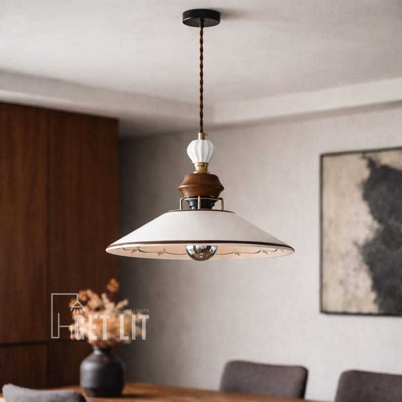 May include: A pendant light fixture with a white, saucer-shaped shade and a decorative floral pattern around the edge. The fixture has a brown and white ceramic accent and a brown twisted cord. The ceiling mount is black.