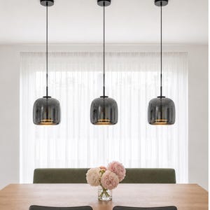 Op de afbeelding: Drie hanglampen met donkergrijze glazen kappen hangen boven een houten eettafel. Elke lamp heeft een zwart snoer en een ronde plafondbevestiging. Op de tafel staat een vaas met roze en witte bloemen.