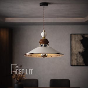 May include: Cream-colored pendant light with a brown and gold trim. The light fixture has a decorative design and a brown cord. The text "GET LIT" is visible in the lower left corner. A stylish lighting fixture for any room.