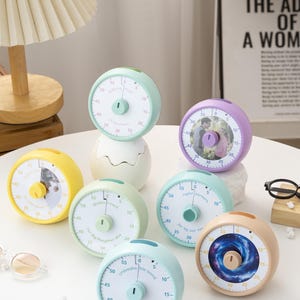 May include: A collection of colorful, round kitchen timers in various pastel shades, each with a white clock face and a central dial. Some timers feature custom text and images, while others have a solid color design. The timers are arranged on a white surface.