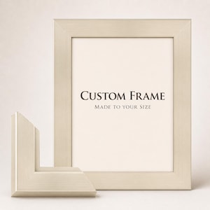 May include: A rectangular picture frame with a brushed silver metallic finish. The frame contains a white insert with the black text "CUSTOM FRAME" and "MADE TO YOUR SIZE". A separate corner sample of the same frame molding is positioned in front.