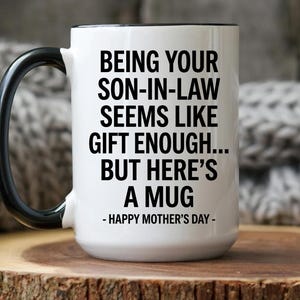 May include: White ceramic mug with a black handle and rim. The mug features the text "BEING YOUR SON-IN-LAW SEEMS LIKE GIFT ENOUGH... BUT HERE'S A MUG - HAPPY MOTHER'S DAY -" in black font. The mug is on a wooden surface.