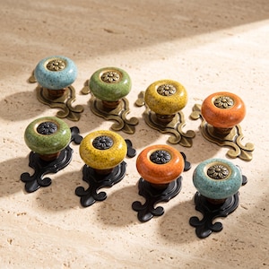 Peut inclure: Un ensemble de huit boutons de placard ronds en céramique bleue, verte, jaune et orange, avec une finition mouchetée et un motif floral métallique central. Les boutons sont fixés sur des plaques métalliques décoratives en bronze ou noir.