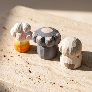 May include: Three hand-carved wooden figurines shaped like animal paws. The left paw is white with orange markings, the center paw is dark gray with pink pads, and the right paw is light beige with brown patches. Each piece has a faceted texture.